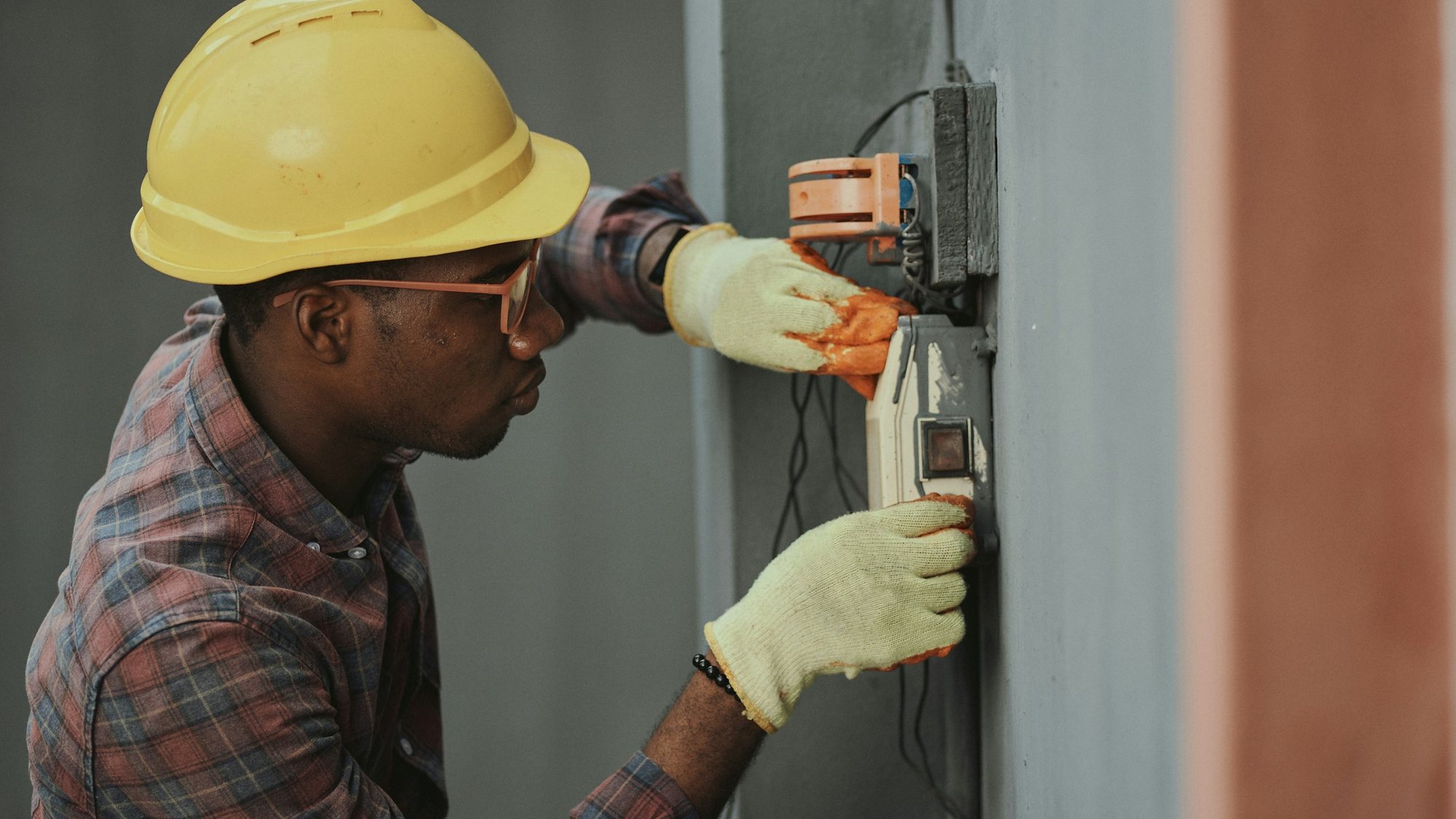 An electrician in a hard hat and safety glasses mounting a device box on a wall.